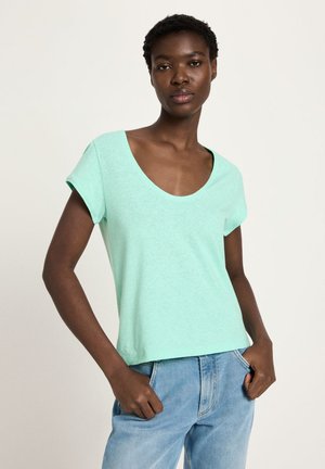 Woman wearing a light blue scoop neck short-sleeve shirt and light wash jeans, standing with hands in pockets against plain background.