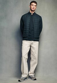 Veste matelassée sombre avec un haut col, t-shirt noir, pantalon crème et baskets grises avec accents noirs, se tenant contre un arrière-plan neutre.