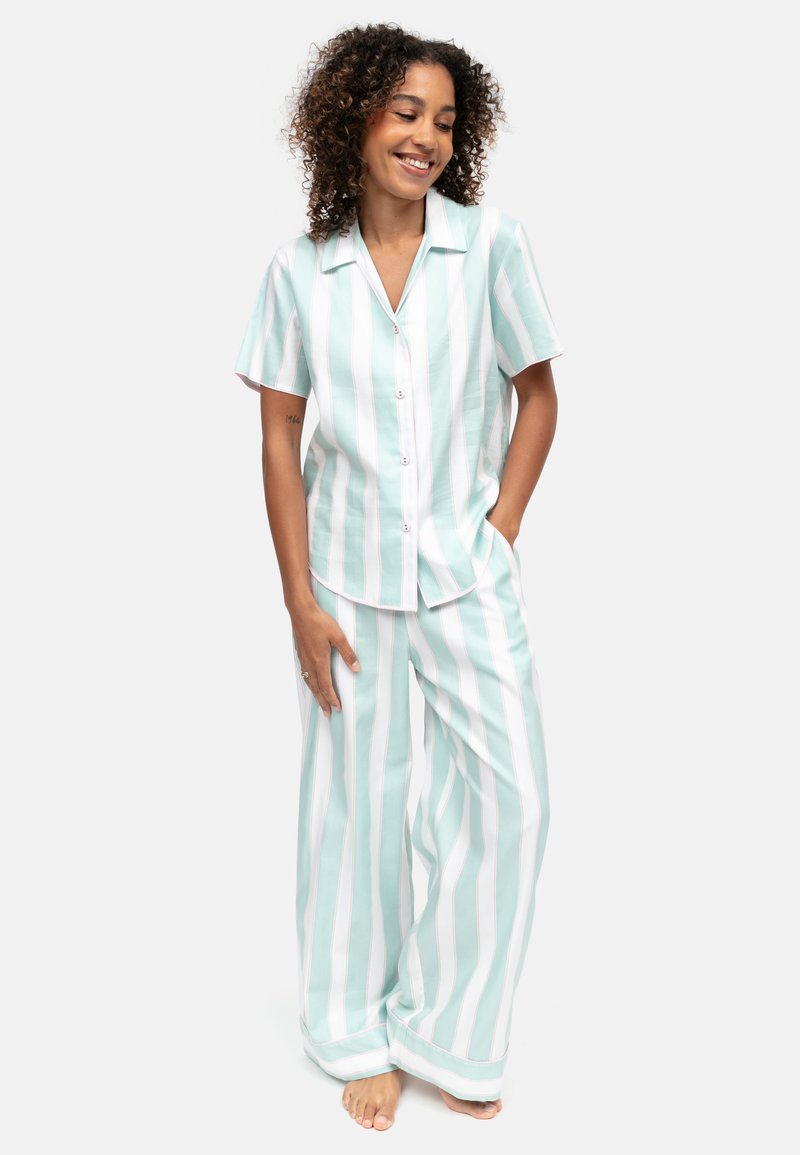 Femme debout pieds nus, souriante, portant un ensemble chemise à manches courtes rayée verticale vert clair et blanc, et pantalon large.