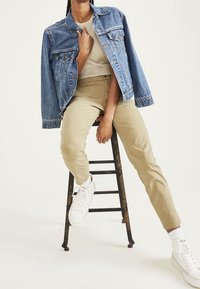 Lichtblauwe spijkerjack met twee borstzakken. Beige slim-fit broeken. Witte sneakers met zichtbare veters en gevoerde tongen, rustend op een kruk.