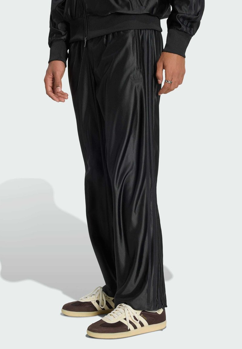 Homme portant un pantalon de survêtement noir avec des bandes latérales et des baskets marron et blanches, debout sur un fond clair.