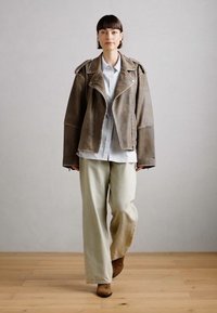 Veste en cuir marron avec un large col, portée sur une chemise bleu clair à boutons. Un pantalon crème ample et des bottes marron complètent le look.