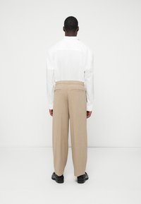 Weißes Hemd mit langen Ärmeln und strukturiertem Kragen, kombiniert mit beigen, weit geschnittenen Hosen mit Gesäßtaschen und subtilen Karomuster. Schwarze Schuhe.