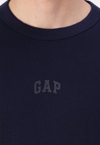 T-shirt côtelé bleu marine avec un col rond et un logo "GAP" gris subtil au centre. Le tissu présente une texture apparente.