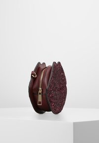 Okrągła torba z burgundowej skóry z błyszczącą czerwoną nakładką, posiada złoty zamek i czarne akcenty, tworząc fakturowany kontrast.
