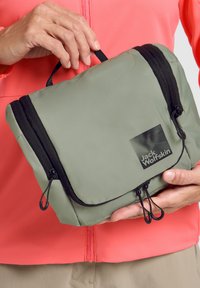Borsa da toiletta verde in nylon con zip nere e anelli regolabili, con logo su un lato. Tenuta da una persona, con uno sfondo di giacca rosa.