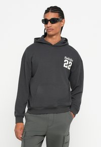 Mladý muž s pletenými vlasy, který má na sobě černé sluneční brýle, tmavě šedou mikinu s nápisem "Hollister 22" a šedé cargo kalhoty, s rukama v kapsách.