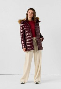 Giacca imbottita bordeaux con texture lucida, cappuccio foderato in pelliccia e chiusura frontale con zip, indossata sopra una maglia rossa e pantaloni beige a gamba larga. Design morbido e isolante.