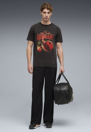 Ung mand iført en sort "Tyskland" grafisk t-shirt, sorte bukser og sneakers, der holder en sort læder duffel taske i den ene hånd.
