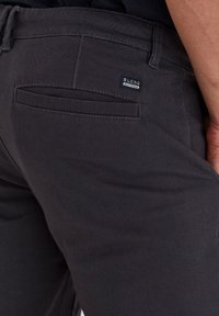 Harmaat puuvilla-sekoitehousut, joissa takataskussa brändäyslabeli. Sileä pinta ja klassinen istuvuus. Yksinkertainen muotoilu ilman näkyviä kuvioita.