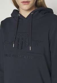 Gros plan d'une personne portant un sweat à capuche bleu foncé avec "GANT 1949" et "American Sportswear" brodés sur le devant.