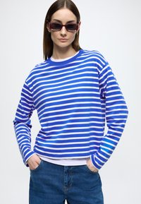 Modrá a bielo pruhovaná košeľa s dlhým rukávom a okrúhlym výstrihom, kombinovaná s vysoko pásovými džínsami. Hladká textúra látky, ležérny strih.