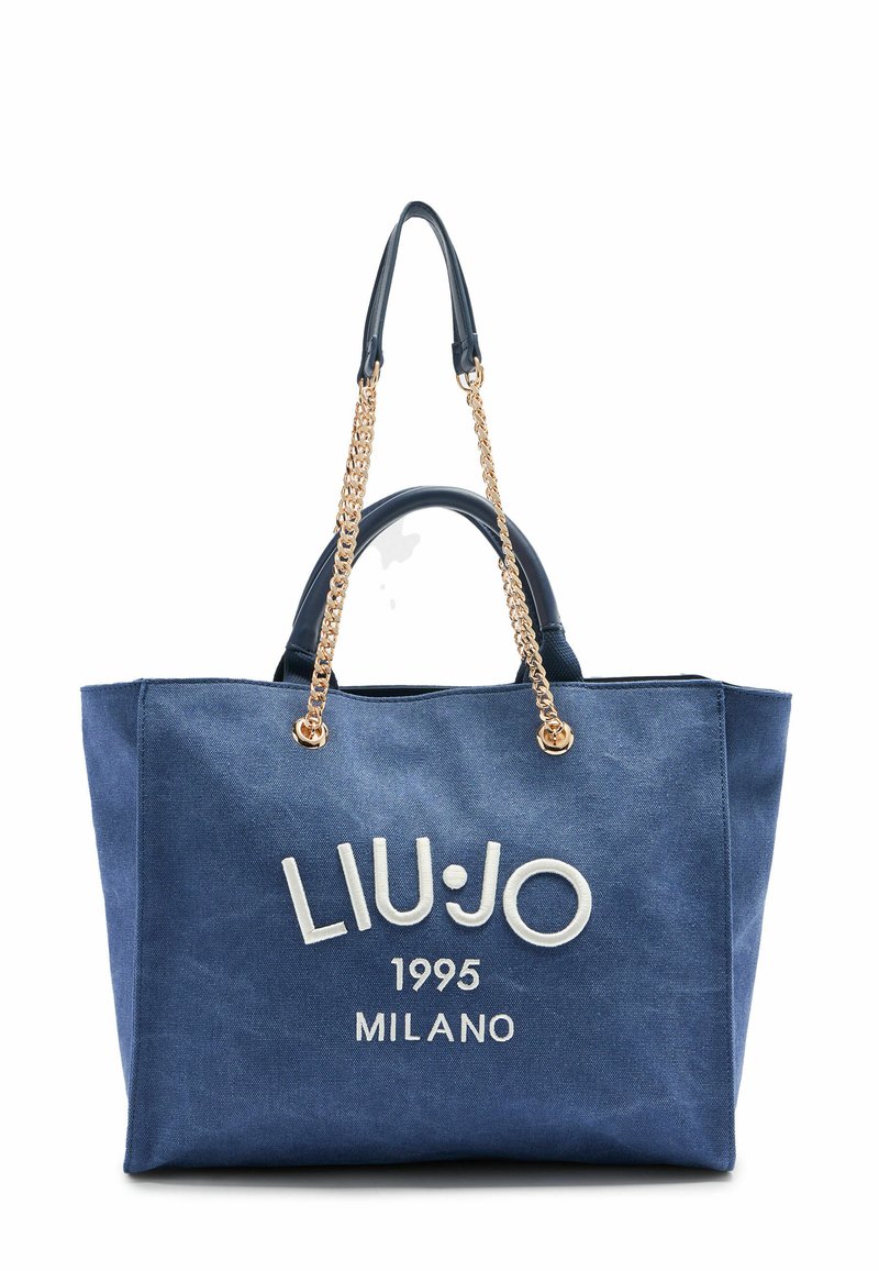 Sac en toile denim bleu avec des anses en chaîne dorée, des poignées en cuir en haut et des détails de logo brodé blanc à l'avant.