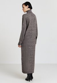 Robe longue et confortable de couleur marron en tricot texturé, avec un col montant et des manches longues, présentant des bords côtelés. Portée avec des bottines noires à la cheville.