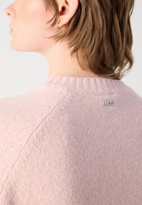 Pull rose clair en tricot texturé, avec un col à côtes et un accent logo argenté à l'arrière près du cou.