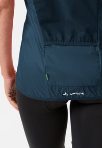 Blå Vaude-jakke med teksturert mesh øvre seksjon, elastisk kant, reflekterende logo og et grønt merkebånd på baksiden.