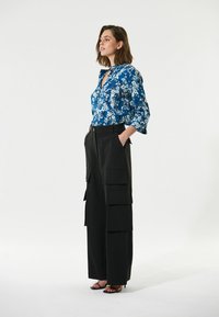 Blaues, blumiges Langarmhemd mit V-Ausschnitt, kombiniert mit schwarzen, weit geschnittenen Hosen mit Cargo-Taschen. Schwarze High Heels.