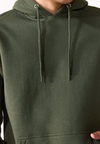 Dunkelgrüne Kapuzenjacke aus weichem, strukturiertem Stoff. Verfügt über eine Kängurutasche, Zugbänder mit Metallspitzen und ein lässiges Design.