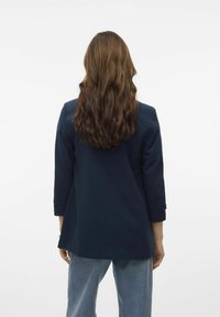 Blazer bleu marine avec une coupe ajustée, manches trois-quarts et texture lisse, vu de dos, associé à un denim clair.