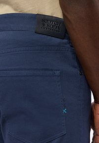 Granatowe spodnie z teksturowanym pasem, z czarną skórzaną metką. Ciekawym szczegółem jest kolorowa haftowana aplikacja w pobliżu kieszeni.