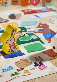 Gul träkanin-token med röd och vit randig sjal, placerad på en färgglad spelbräda med olika illustrerade objekt och djur.