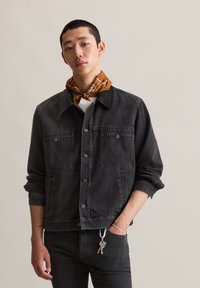 Zwarte spijkerjack met een klassieke kraag, voorzakken en zilveren knoopsluitingen. Model draagt een bruin patroon sjaal en slim fit zwarte broek.