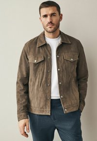 Bruine suède jas met een kraag, sluiting met knopen aan de voorkant en twee borstzakken. Draagt over een witte t-shirt en gecombineerd met donkere broek.