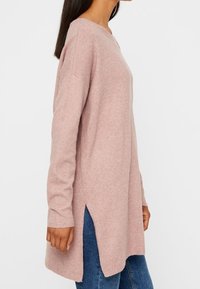 Eine Frau trägt einen langen, hellrosa Strickpullover mit seitlichen Schlitzen über blauen Jeans, seitlich vor einem schlichten Hintergrund gezeigt.