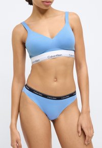 Ανοιχτό μπλε bralette με ντεκολτέ σε σχήμα V και φαρδιά λευκή ταινία με το λογότυπο «Calvin Klein». Συνοδευτικό μπλε στρινγκ με μαύρη μέση.