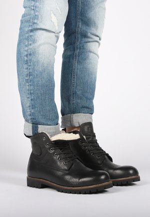 Botas de tobillo de cuero negro con acabado texturizado, diseño con cordones, forro de piel de pelaje y suela resistente. Combinadas con jeans azul claro arremangados.