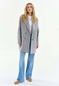 Žena s dlhými blond vlasmi, oblečená v šedom kabáte s rybej kosťou, bielej košeli, modrých rozšírených džínsoch a béžových slip-on topánkach, stojaca pred bielym pozadím.