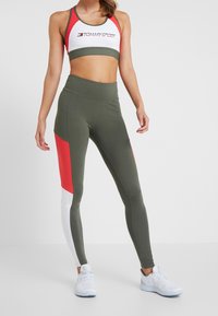 Sport Crop-Top in Weiß mit roten Akzenten und Logo; olivgrüne, hoch taillierte Leggings mit roten und weißen Farbblöcken. Das Material scheint dehnbar zu sein.