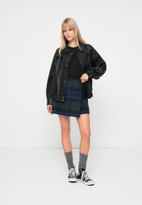 Črna usnjena jakna, črna srajca s gumbi, modra in zelena karo mini krilo, sivi kolenski nogavici in črne superge. Model stoji ob enostavni ozadju.