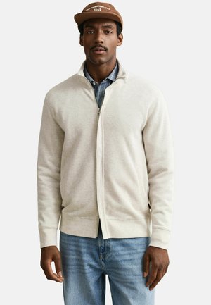 Ein Mann trägt eine beige Reißverschlussjacke über einem blauen Hemd mit Knöpfen, blaue Jeans und eine braune Kappe mit weißem Schriftzug.