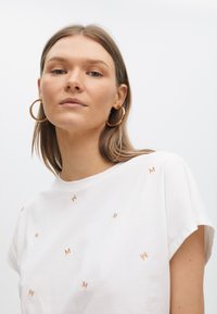 T-shirt en coton blanc avec de petites lettres "M" brodées en marron, dispersées sur tout le tissu, manches courtes et encolure ronde.