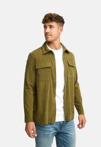 Olivengrøn kordfløjljakke med to klaplommer, lynlås og krave, iført over en hvid T-shirt, parret med blå jeans.