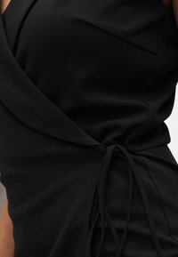 Robe portefeuille noire avec un nœud décoratif sur le côté, portée sur le torse d'une personne avec bras et épaule visibles.