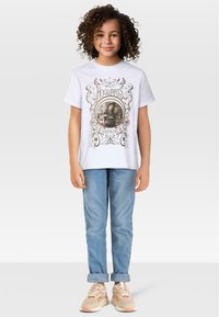 Biała koszulka z graficznym nadrukiem przedstawiającym "Powrót do Hogwartu" oraz wizerunek Hermiony, w połączeniu z jasnoniebieskimi dżinsami i beżowymi trampkami.