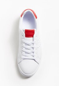 Baskets blanches avec un accent rouge au talon, design à lacets, matériau synthétique lisse, logo embossé sur la languette, et semelle texturée.