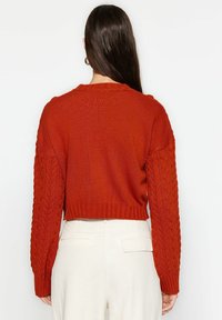 Femme aux longs cheveux foncés portant un pull tricoté couleur rouille avec des manches torsadées et un pantalon blanc taille haute, vue de dos.