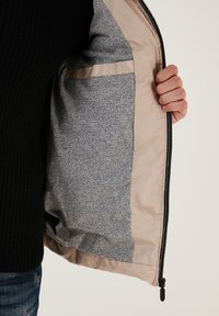 Letvægtsjakke med en beige indvendig, som har et glat gråt stoffor og en sidelomme. Udsiden har en lynlåslukning.