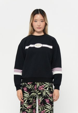 Sweat-shirt noir avec le logo "ROXY", rayures blanches et lavande sur les manches, poignets côtelés et coupe décontractée, associé à un pantalon à motifs floraux.