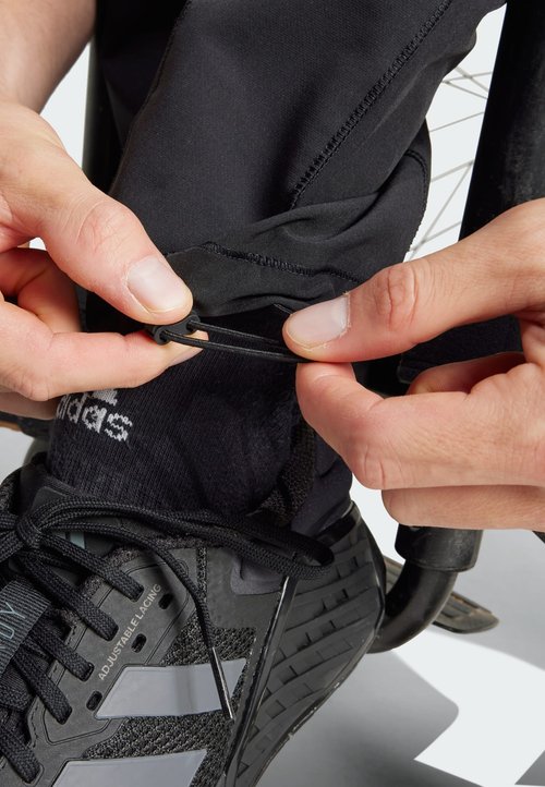 Pantalones negros ajustables con puños elásticos y manos tirando de un cordón; zapatos deportivos negros con suelas texturizadas.