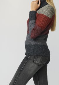 Pull en maille à col roulé avec des rayures horizontales en gris foncé, rouille et gris clair. Poignets et ourlet côtelés. Porté avec un jean noir.
