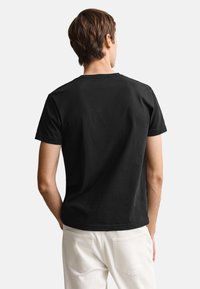 Muž s hnedými vlasmi, ktorý má na sebe jednofarebné čierne tričko s krátkym rukávom a biele nohavice, stojí chrbtom ku kamere pred bielym pozadím.