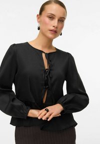Juoda tekstūruota bluzonė su pūstomis rankovėmis, priekiniais užsegimais ir surišta talija. Modelis dėvi juodą žiedą ir ryškias auskarus.