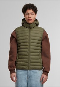 Olivenfarget puffervest med horisontale sømmer, høy krage og full glidelås. Brun langermet skjorte under, kombinert med lyseblå denim.