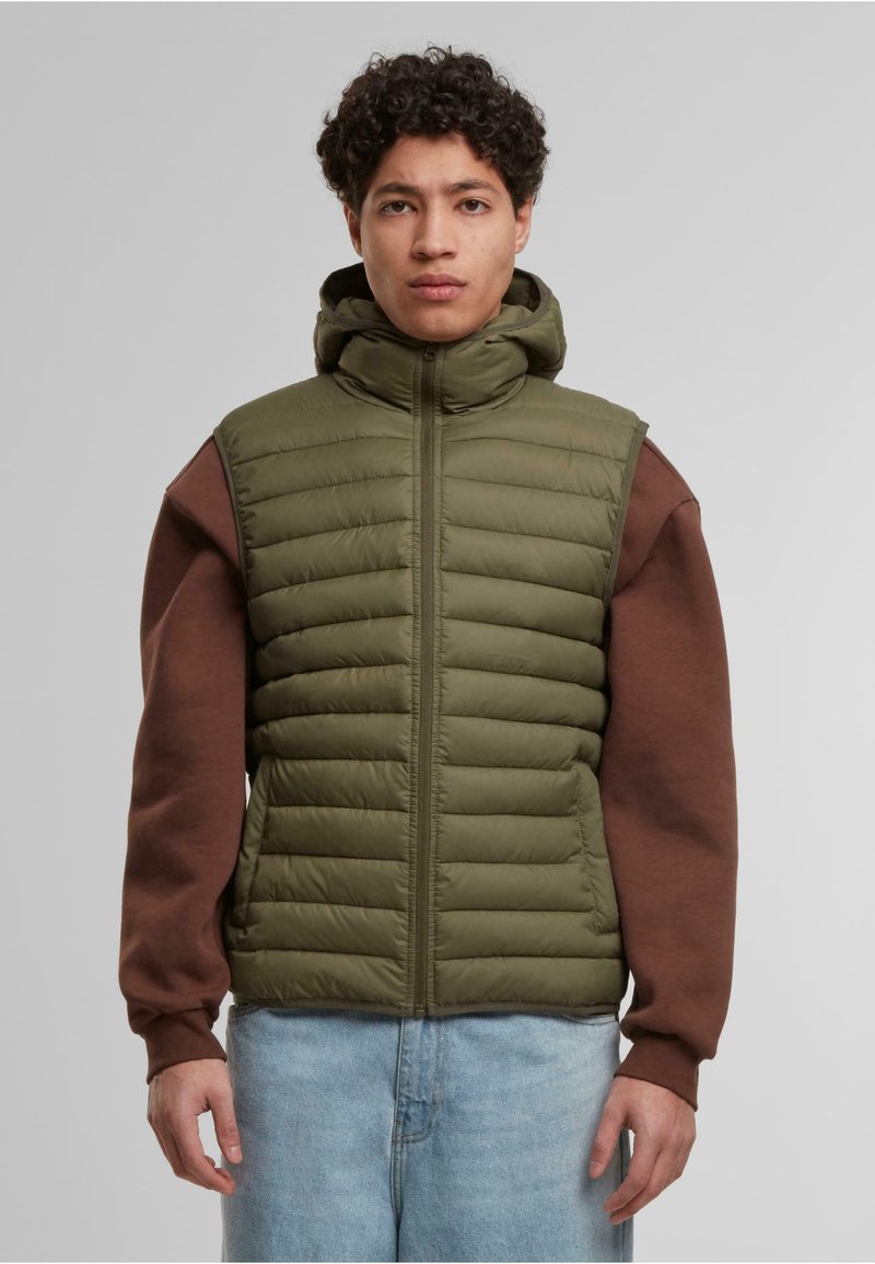 Olivenfarget puffervest med horisontale sømmer, høy krage og full glidelås. Brun langermet skjorte under, kombinert med lyseblå denim.