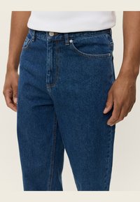 Donkerblauwe denim jeans met een klassieke snit, voorzien van oranje stiksels, een knoopsluiting en voorkezen. Materiaal heeft een gladde textuur.
