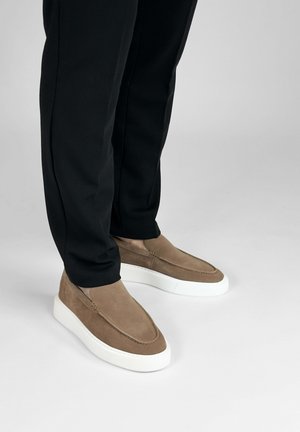 Voeten met bruine suède instapsneakers met witte zolen en een zwarte broek staand op een lichtgrijze vloer.